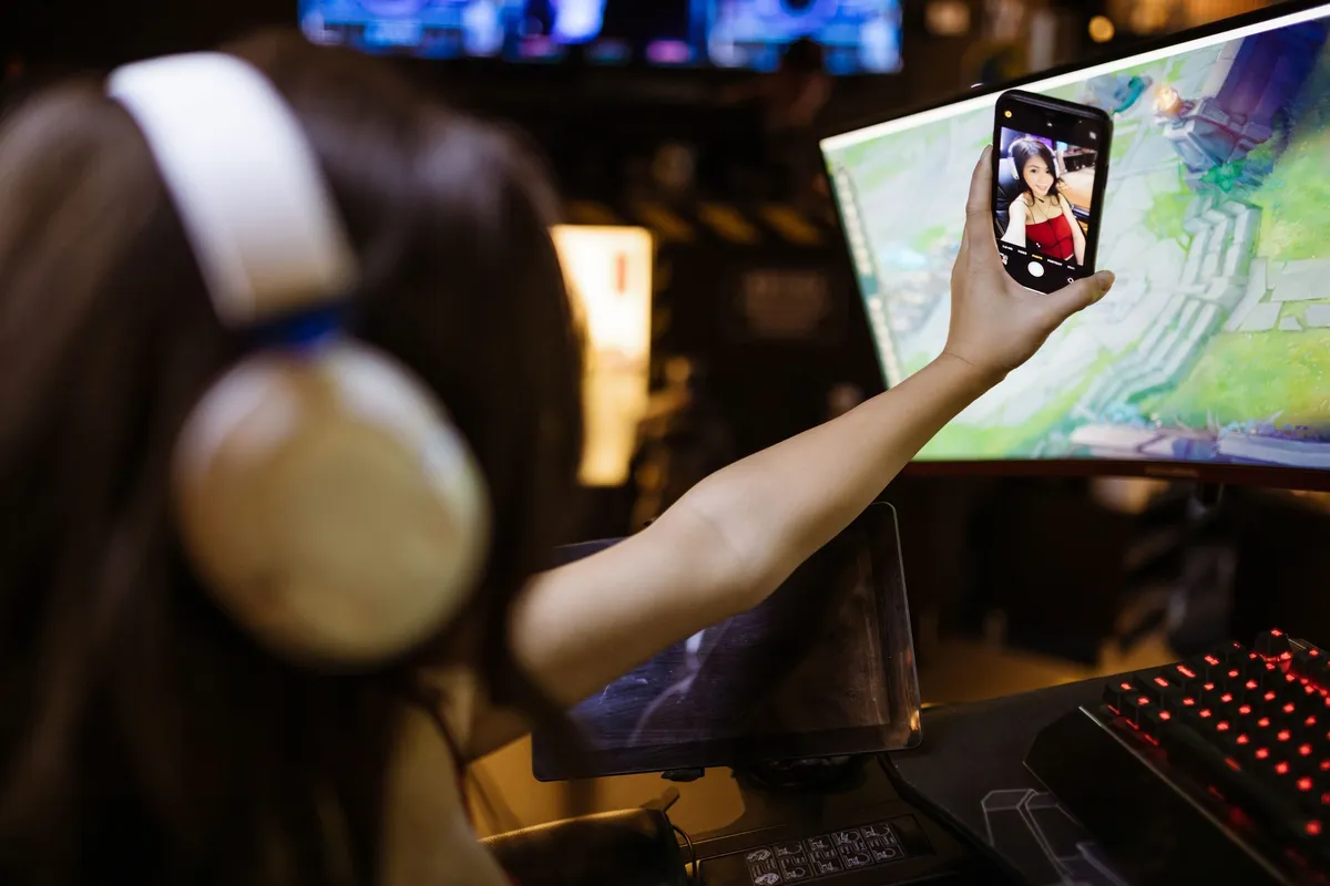 A player in headphones holding up a phone to film herself in front of a game on a monitor