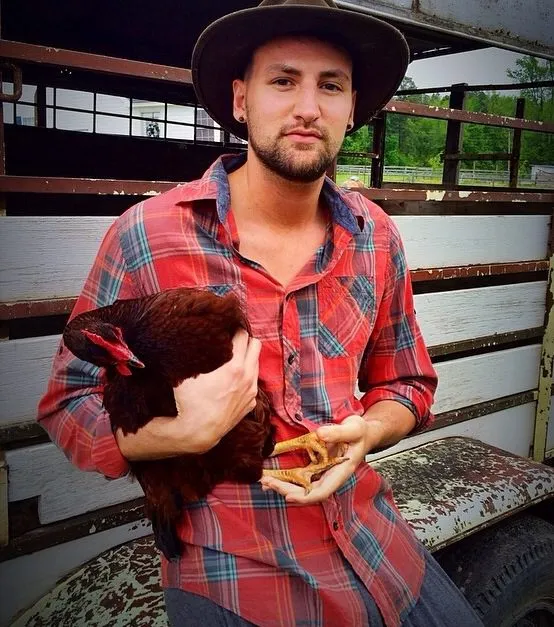 Alx James in a plaid shirt and brimmed hat holding a brown hen inside a livestock trailer