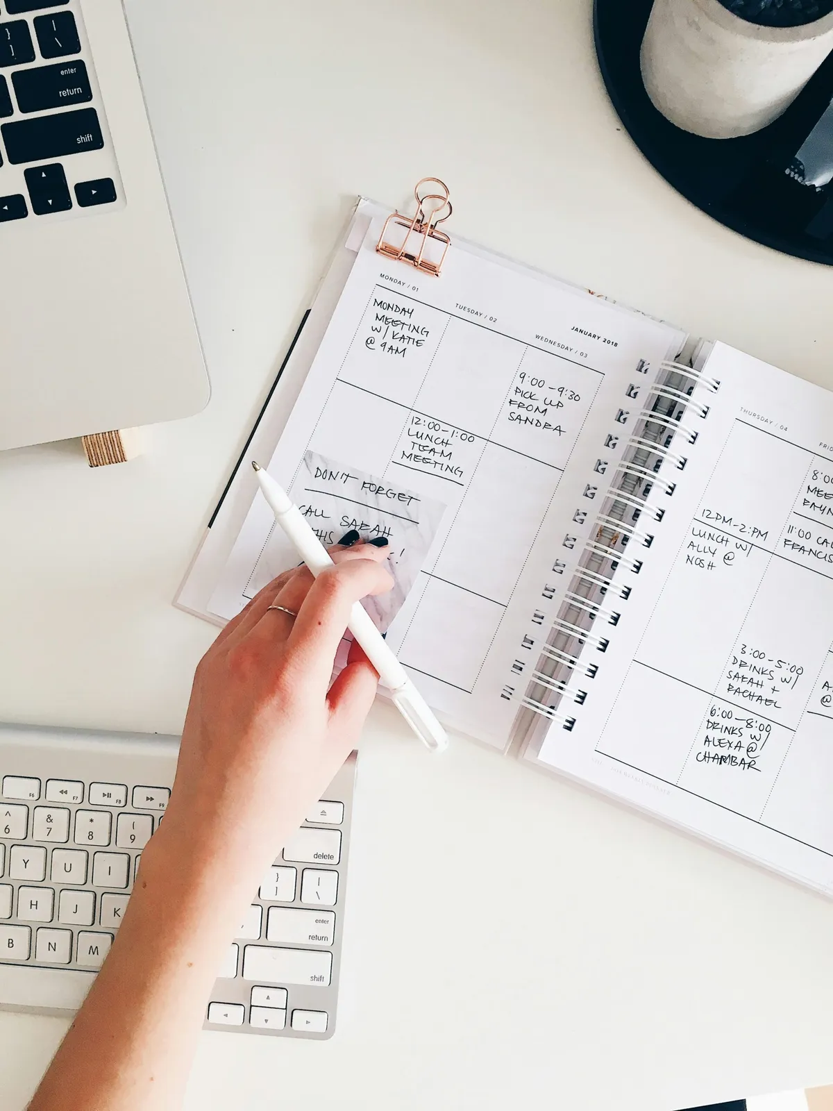 Hand writing weekly appointments in an open paper planner beside a laptop and keyboard on a white desk