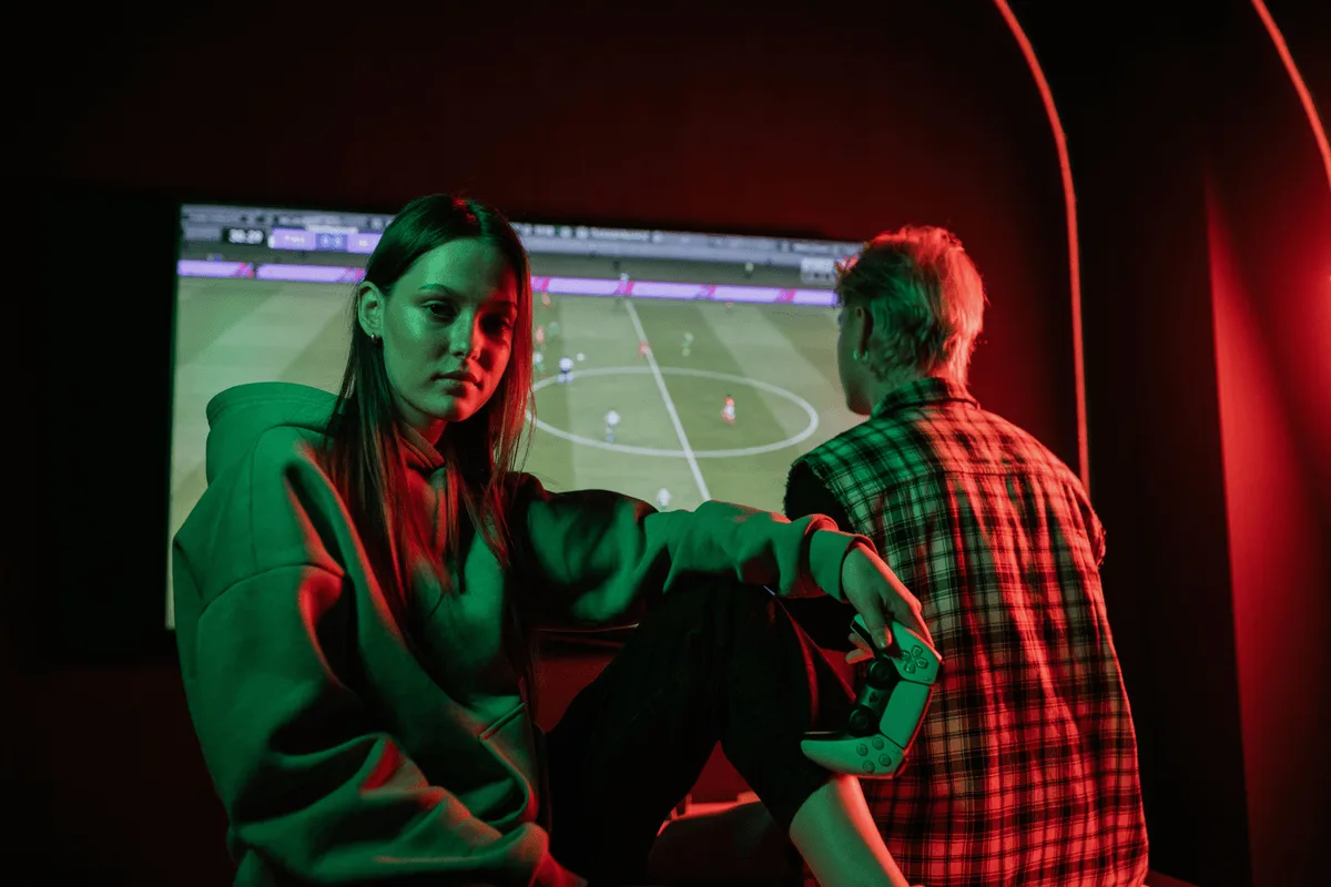 Two players lit in green and red sitting in front of a large screen showing a football game
