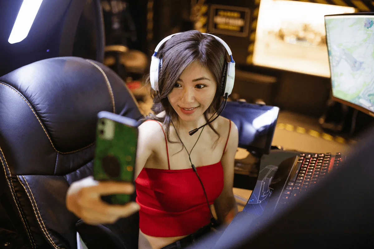 A gamer in a red top and white headset holding up a phone to film herself at a lit gaming rig