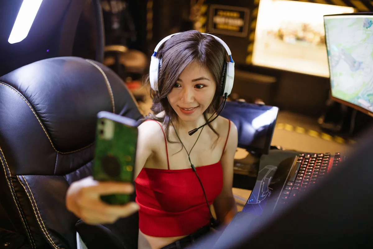 A gamer in a red top and white headset holding up a phone to film herself at a lit gaming rig