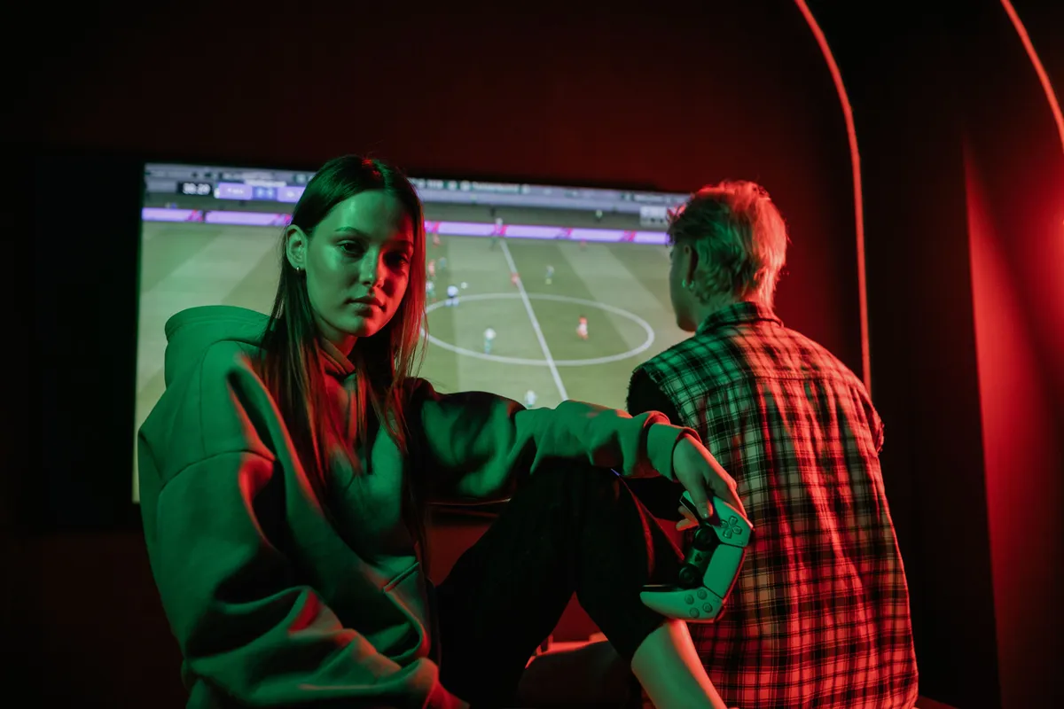 Two players lit in green and red sitting in front of a large screen showing a football game
