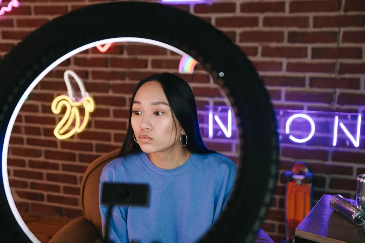 A creator framed by a ring light, filming against a brick wall with neon signs behind her