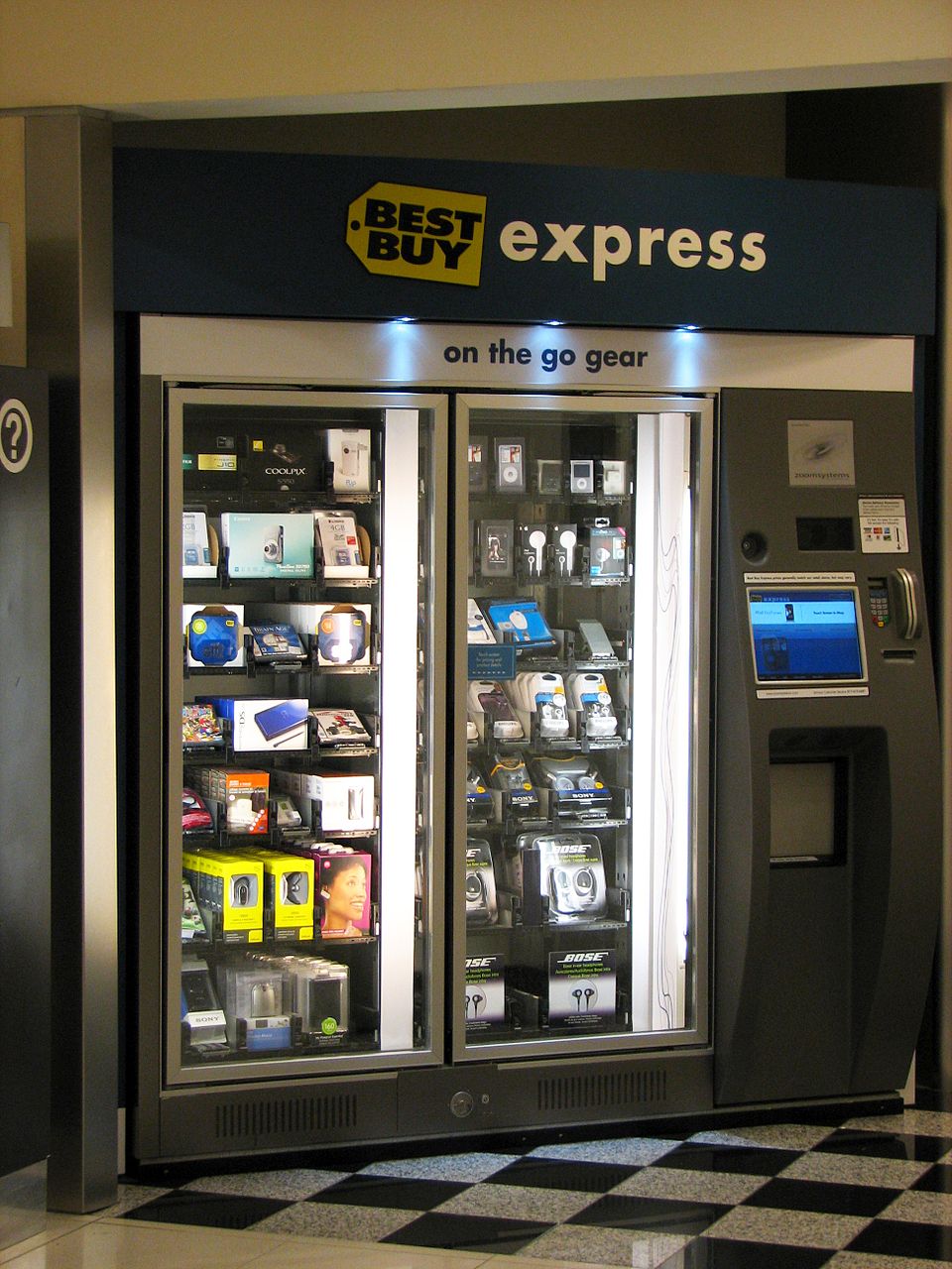 A Best Buy Express vending machine by Zoom Systems at an airport terminal, stocked with electronics.