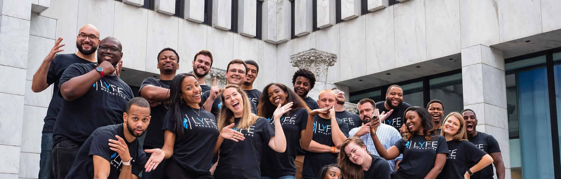 A diverse and energetic group of approximately twenty professionals from the Lyfe Marketing team poses for a wide-angle group photo outdoors. The team members are mostly wearing matching black t-shirts featuring the Lyfe Marketing logo and are seen smiling, laughing, and making playful gestures, creating a vibrant and collaborative atmosphere. They are positioned against the backdrop of a modern white stone building with large glass windows and architectural pillars.