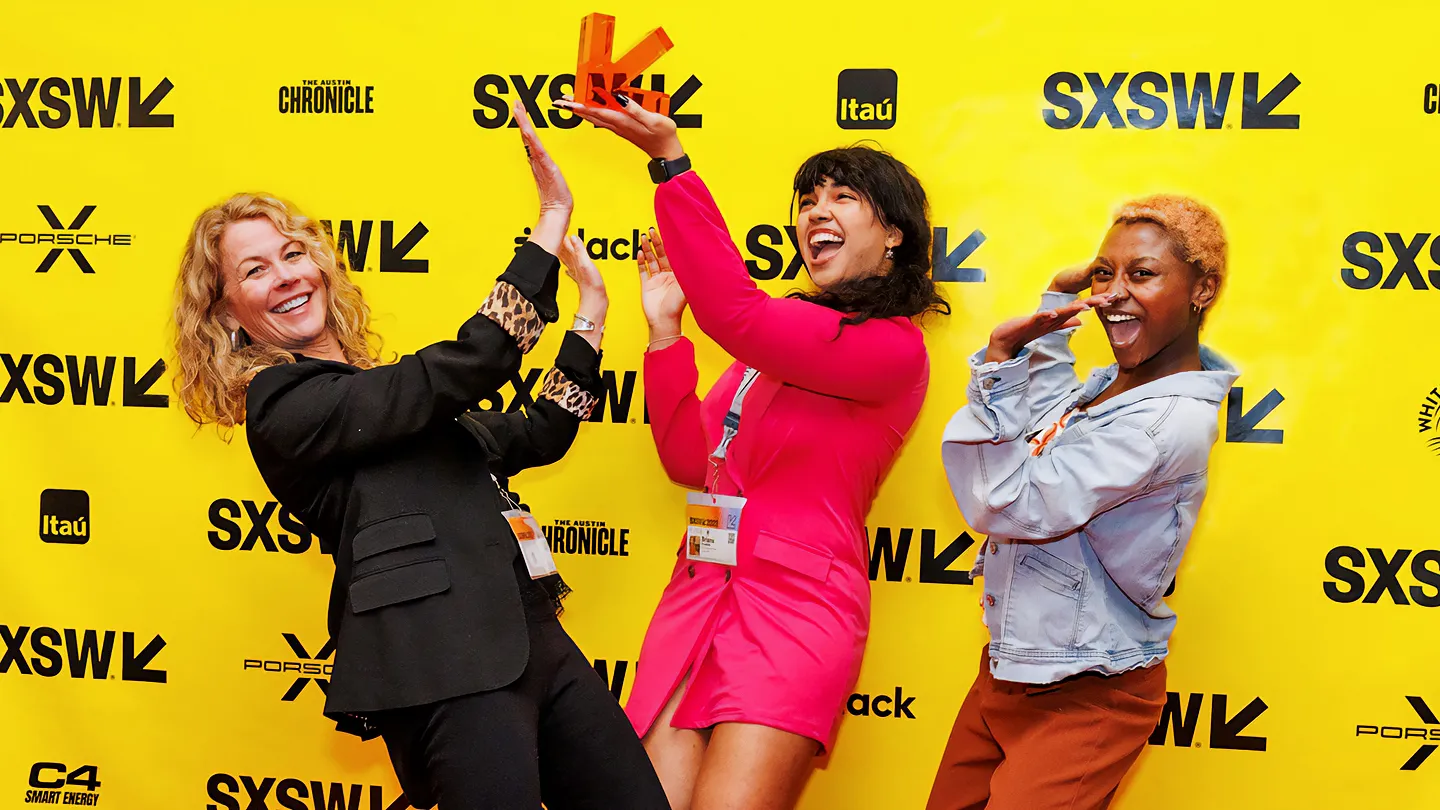 Three women celebrate joyfully at the SXSW 2023 Innovation Awards, posing in front of a bright yellow backdrop branded with logos for SXSW, Porsche, and The Austin Chronicle. On the left, a woman in a black blazer and black pants smiles with her hands raised. In the center, a woman in a hot pink blazer mini dress, which is unlined and suitable for a cocktail or party atmosphere, laughs while holding up a small orange award. To the right, a third woman in a light denim jacket and rust-colored pants strikes a festive pose to complete the scene.